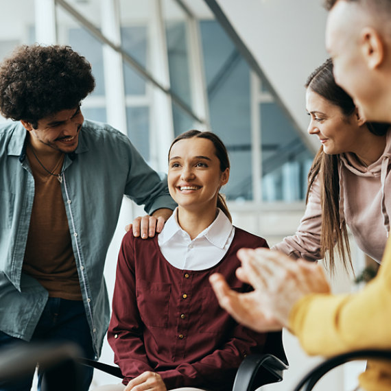 A woman in a wheelchair is congratulated by several colleagues in a modern office.