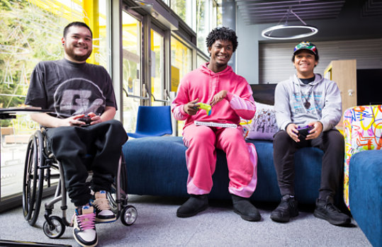 Logo of Xbox on the left and two youths, one with an adaptive controller, play video games.