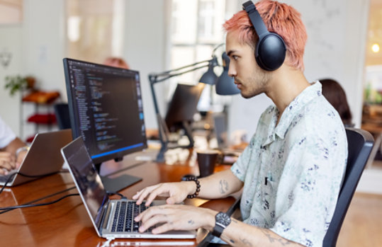 Un uomo che indossa delle cuffie e lavora sul portatile in ufficio e logo di Microsoft Visual Studio a destra.