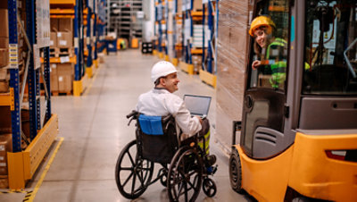 Man in a wheelchair using a computer and a young woman in a forklift smile while working in a factory.