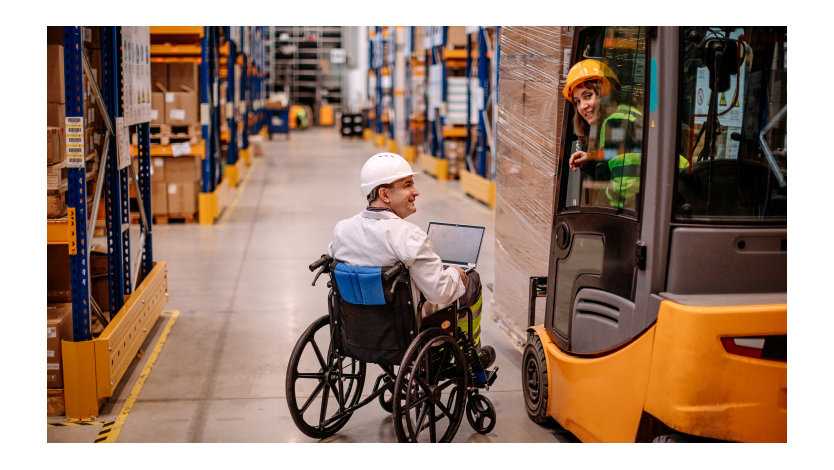Un hombre en silla de ruedas usando un ordenador y una mujer joven en una carretilla elevadora sonríen mientras trabajan en una fábrica.