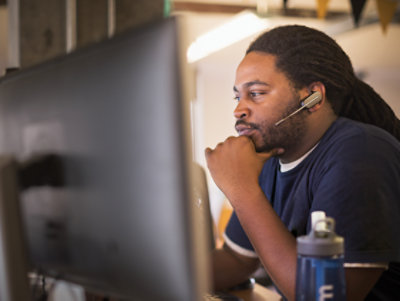 A man with a headset works intently at a computer in an office setting, hand on chin, deep in thought.