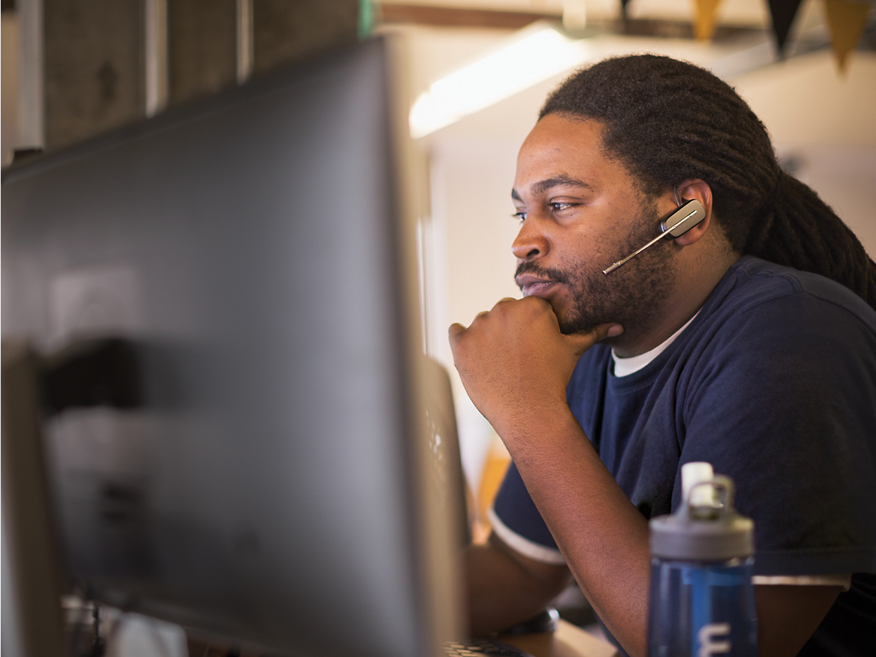 Seorang pria menggunakan headset bekerja dengan fokus menggunakan komputer di dalam lingkungan kantor, tangan di dagu, sedang berpikir mendalam.