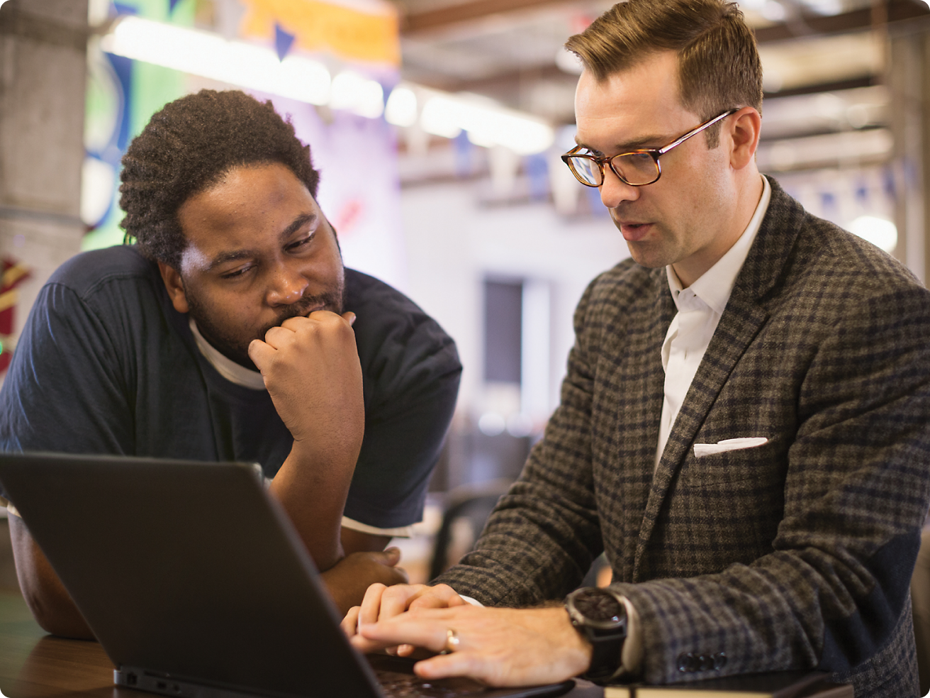 Dois homens, um usando óculos, revisam conteúdo em um laptop juntos em um ambiente de escritório.