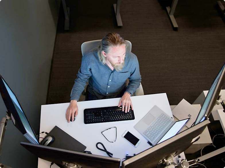 Un bărbat stând la o stație de lucru de birou, utilizând o tastatură cu mai multe monitoare și un laptop pe birou.