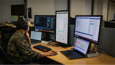 Une femme en vêtements de camouflage est assise à un bureau devant deux écrans.