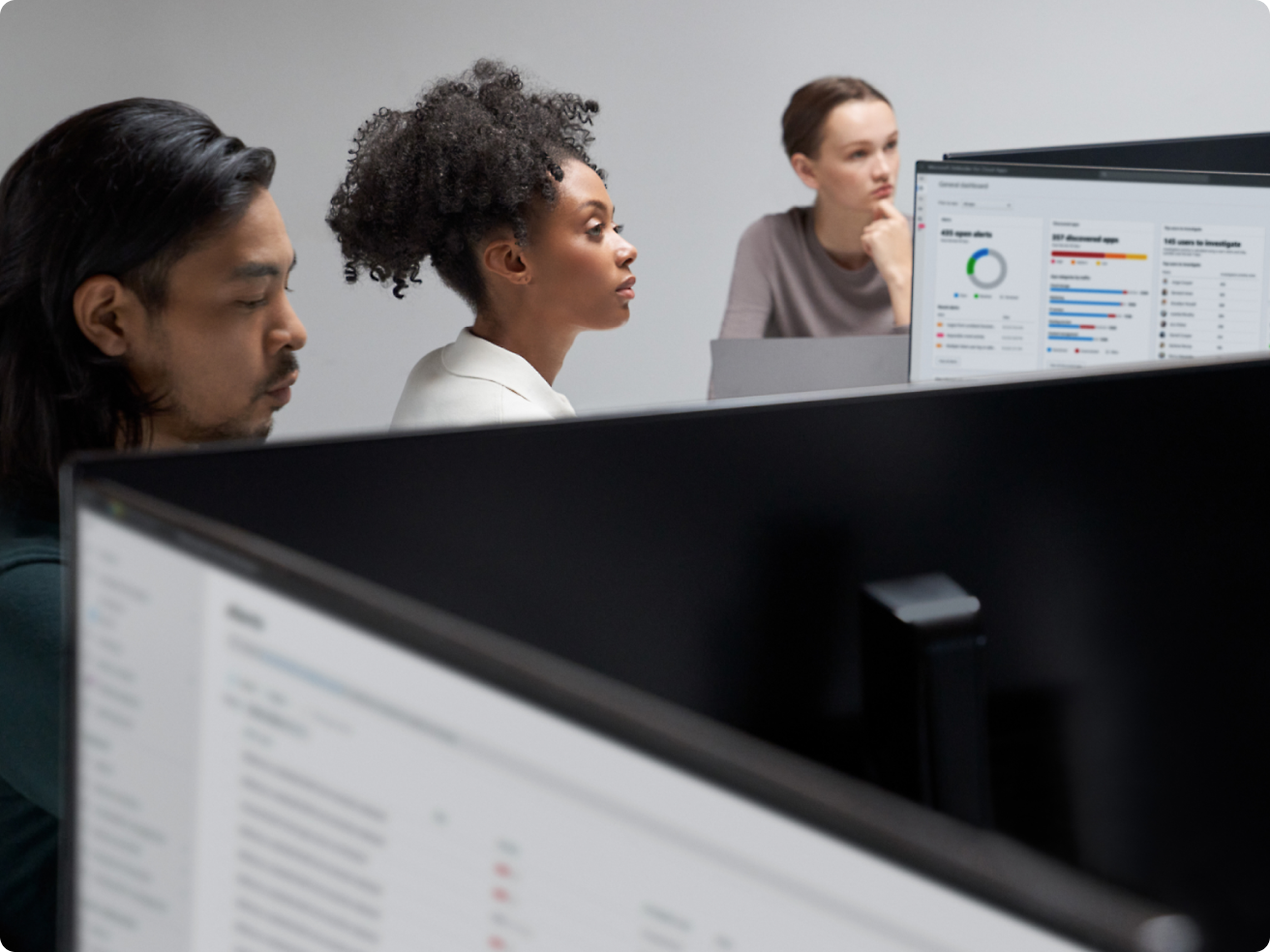 Trys biuro darbuotojai, susitelkę į kompiuterių ekranus, kuriuose vaizduojami grafikai ir duomenys modernioje biuro aplinkoje.