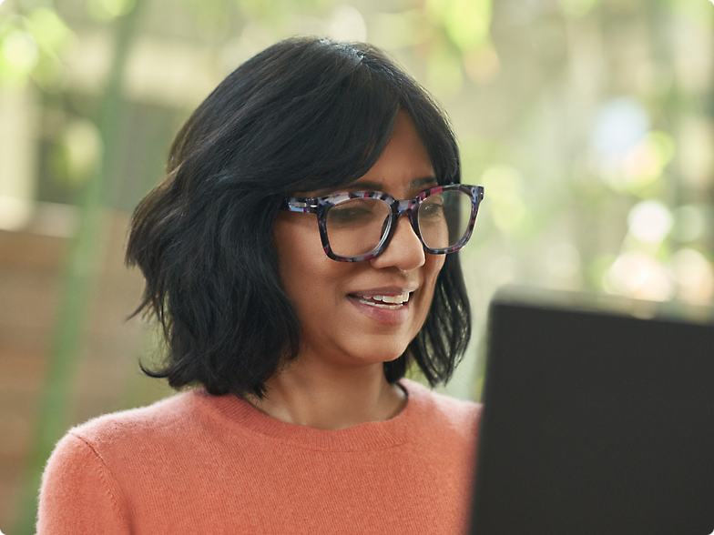 Uśmiechnięta kobieta w okularach ubrana w sweter brzoskwiniowy, spoglądająca na ekran laptopa w nasłonecznionym pomieszczeniu.