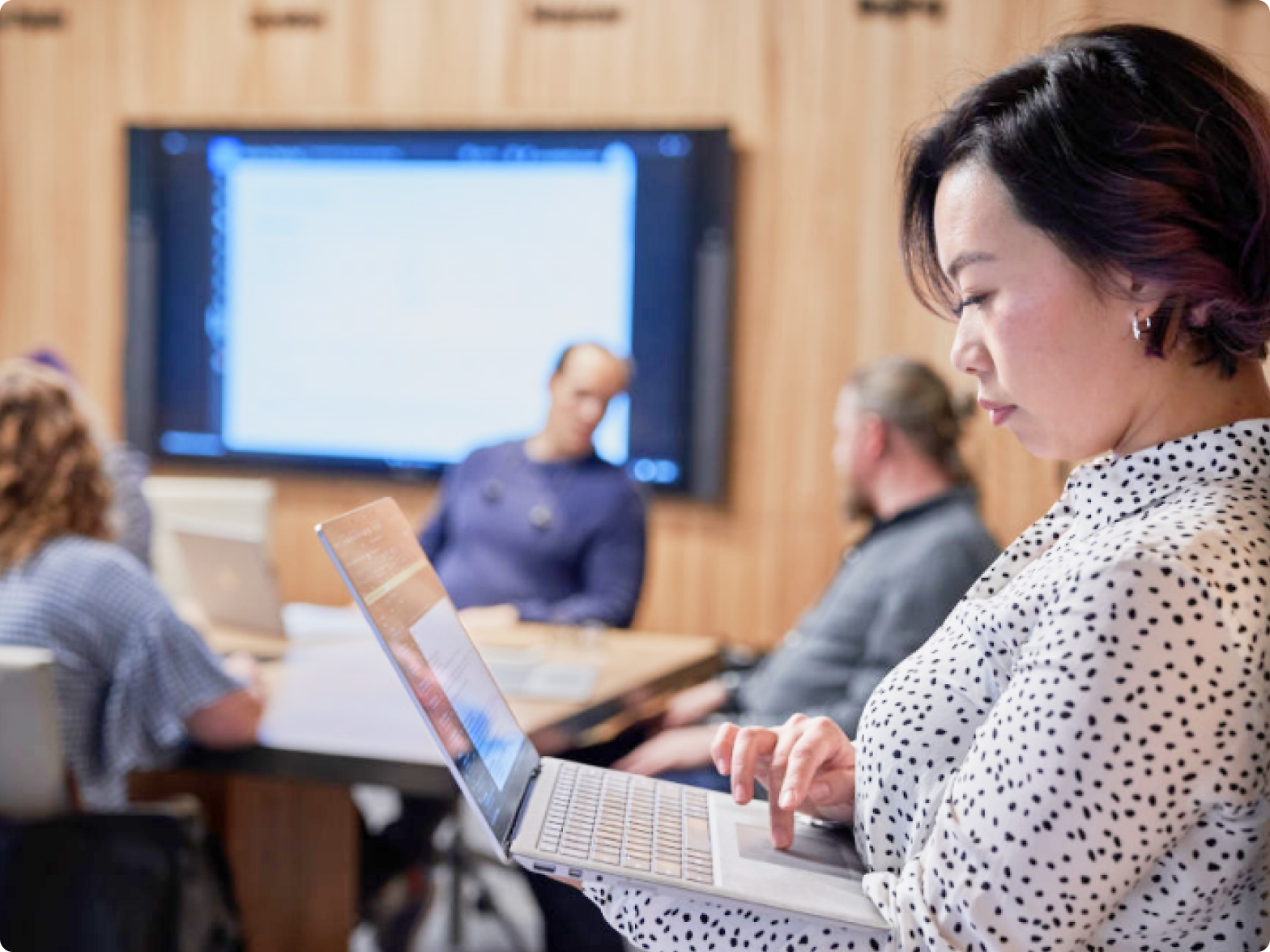Uma mulher a utilizar um portátil numa sala de conferências com três outros colegas e um ecrã de apresentação em segundo plano.
