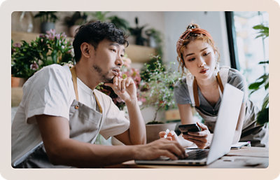 Két kötényes ember, az egyik laptopot használ, a másik telefont tart, és a képernyőt nézi