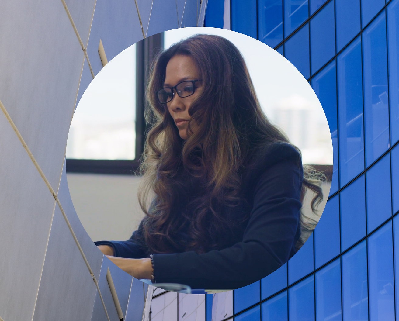 Uma mulher a trabalhar no seu portátil com óculos e uma imagem de fundo de um edifício de vidro