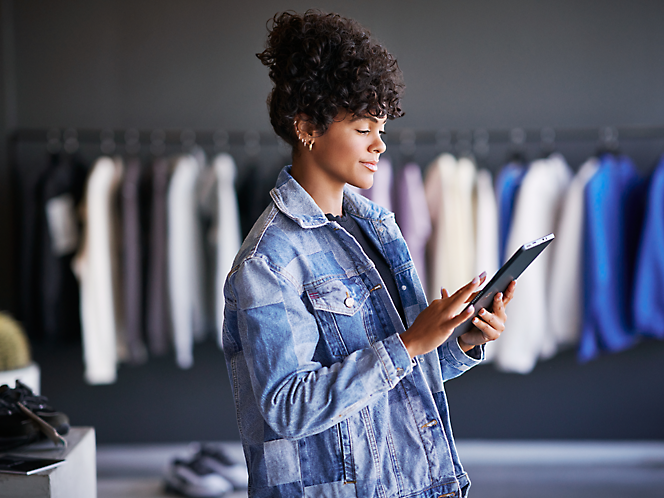 A young woman in a denim jacket using a tablet in a clothing store with racks of clothes in the background.