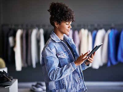 Eine Frau in einer Jeansjacke, die ein Tablet in einem Bekleidungsgeschäft nutzt.