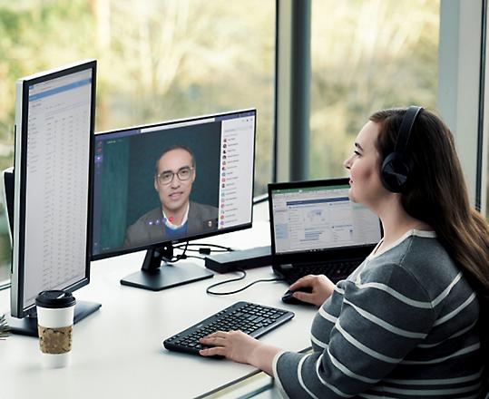 A person participating in a video meeting from their home desk with multiple screens and over the ear headphones on 