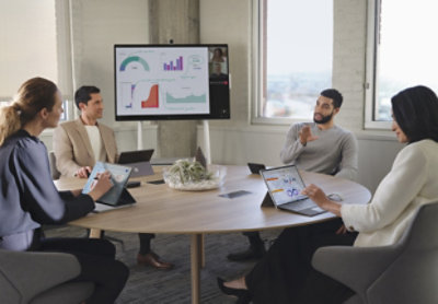 Vier Personen, die eine Besprechung in einem Konferenzraum abhalten, während im Hintergrund ein Teams-Anruf angezeigt wird