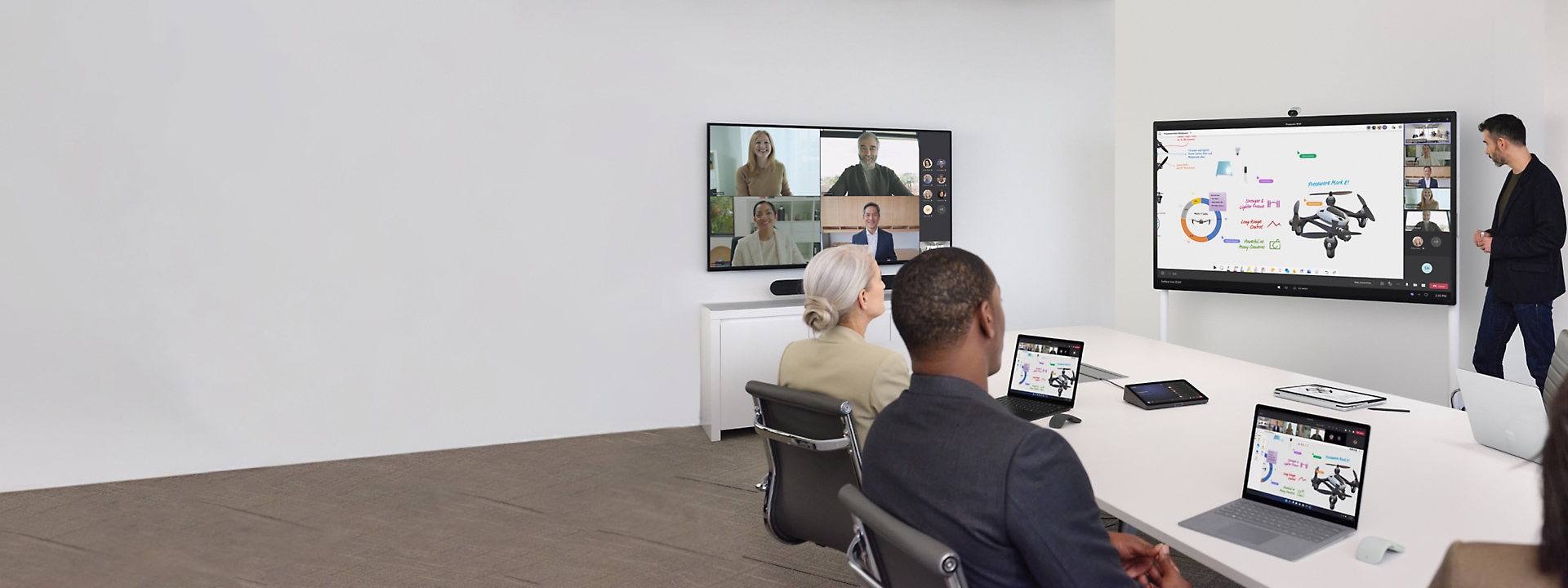 Gente en una sala de conferencias teniendo una reunión con una televisión que muestra una videollamada de Teams y otra que muestra una presentación.
