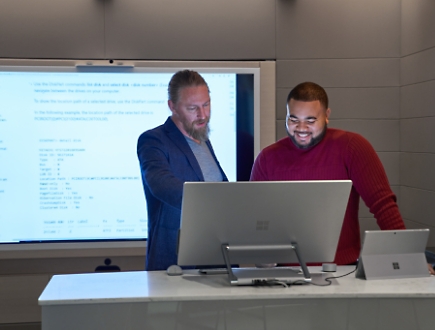 Два человека работают вместе за настольным компьютером перед большим экраном, на котором отображается код. 