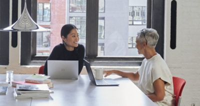 Twee personen die met hun laptops in een vergaderruimte zitten en een gesprek voeren