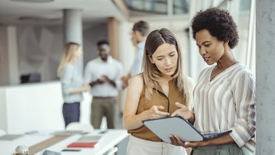 Two people standing together in an office looking at a laptop while a small group of employees have a conversation in the background 