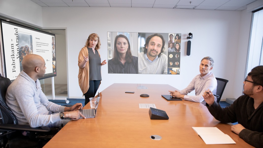 A meeting with four people in a conference room and more people dialed in through a Teams video call being projected on a wall.