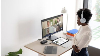 A person standing in a home office wearing over-the-ear headphones and participating in a Teams video call on a monitor on their desk.