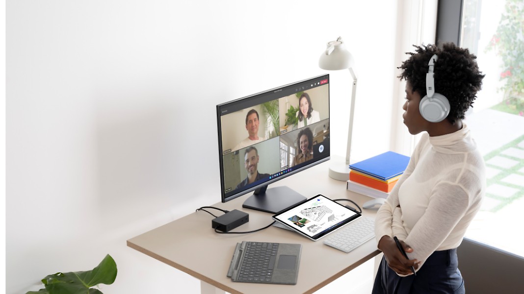 Oseba stoji v domači pisarni z naglavnimi slušalkami in sodeluje v videoklicu v aplikaciji Teams na monitorju na svoji mizi.