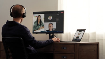 A person sitting in their home office with a laptop and a desktop monitor participating in a Teams call with three other participants.