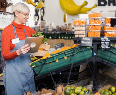 A person working in a grocery store holding a tablet and looking at a bin of oranges