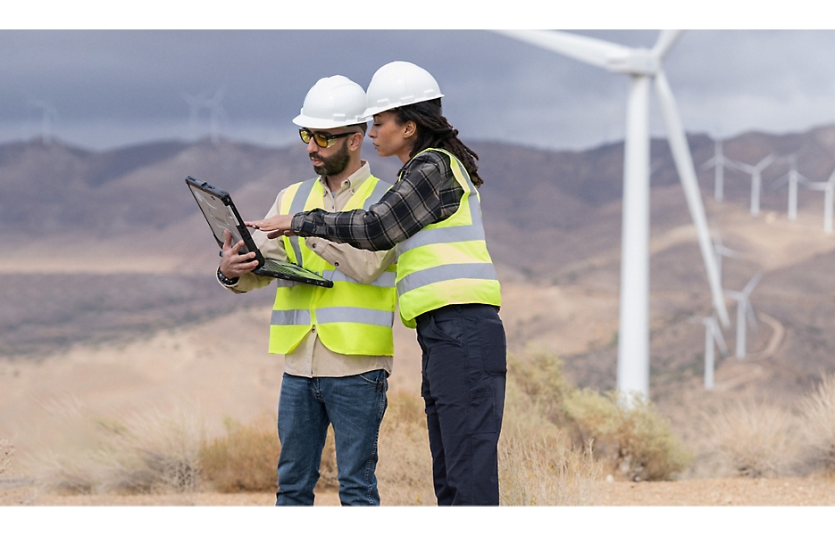 Duas pessoas usando capacetes e coletes de segurança em pé em um campo com turbinas eólicas olhando para um laptop