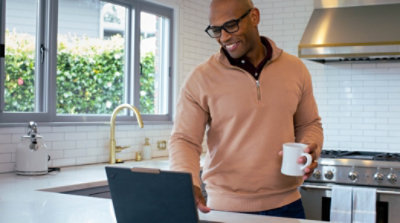 A person standing at a kitchen counter holding a coffee mug and using a laptop