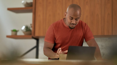 Persona, kas sniedzas pēc kafijas krūzes un izmanto klēpjdatoru savā virtuvē