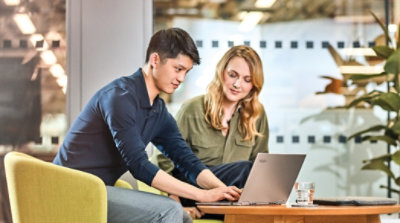 Divi cilvēki sēž viens otram blakus klubkrēslos, viena persona strādā klēpjdatorā, bet otra skatās ekrānā