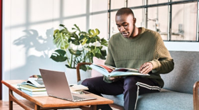 A student sitting on a couch reading a textbook with a laptop open on the table in front of them
