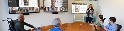 A meeting being held in a conference room with many members joining remotely through a Teams video call being displayed on multiple screens in the room.