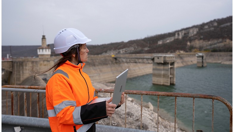 A construction worker holding a tablet in front of a dam.