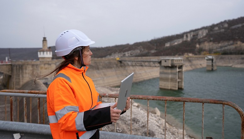 A construction worker holding a tablet in front of a dam.
