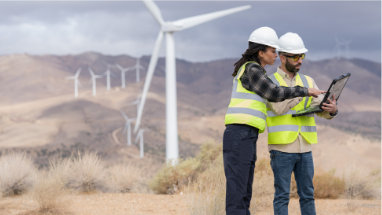 Due persone che indossano indumenti protettivi davanti a turbine eoliche e guardano un computer portatile.