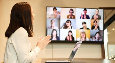 A person in a meeting room speaking to a group of people over a video call being displayed on a large screen on the wall