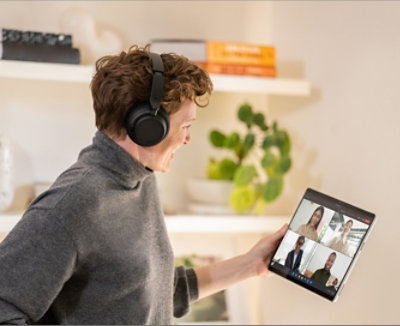 A person wearing over the ear headphones and holding a tablet displaying a Teams video call