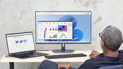 Persona, kas strādā pie sava rakstāmgalda ar klēpjdatoru un darbvirsmas monitoru, kurā darbojas attālais dators