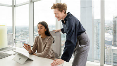 A person leaning over a coworker sitting at their desk and having a conversation over a tablet