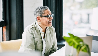 A woman wearing glasses is laughing in front of a laptop.