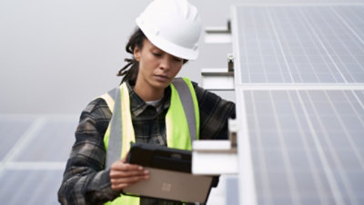 Una persona con un casco y un chaleco de seguridad trabaja en un panel solar y consulta una tableta