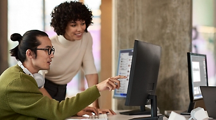 A person sitting at a desk pointing at data on a desktop monitor and speaking to a coworker leaning over their shoulder