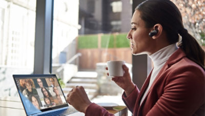 A person wearing an in-ear Bluetooth device holding a cup of coffee and participating in a Teams video call on a laptop with 8 other participants.
