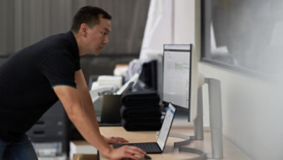 A person leaning over a desk using a laptop connected to a desktop monitor