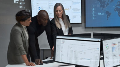 Three people standing over a desk looking at data being displayed on a desktop monitor