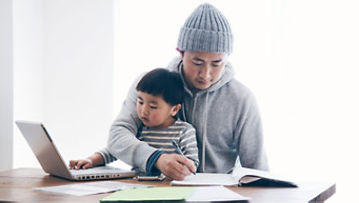 A man writes in a notebook while his kid sits on his lap and plays with a laptop