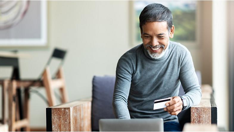 Hymyilevä henkilö pitää luottokorttiaan kädessä ja syöttää sen tietoja kannettavalle tietokoneelleen. 