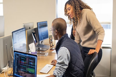 Three people having a conversation at a desk with multiple screens.